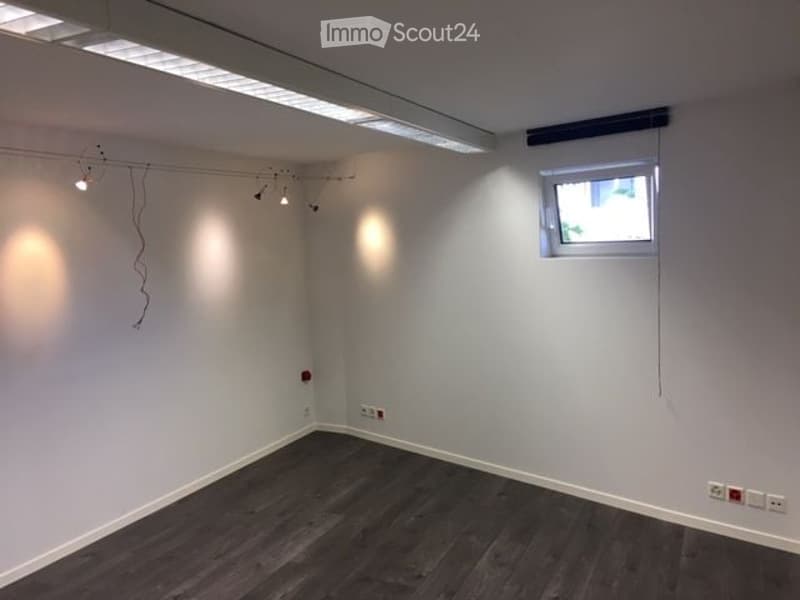 Empty room with white walls, dark wooden flooring, and a small window with blinds. Exposed electrical wiring and outlets are visible on the walls.