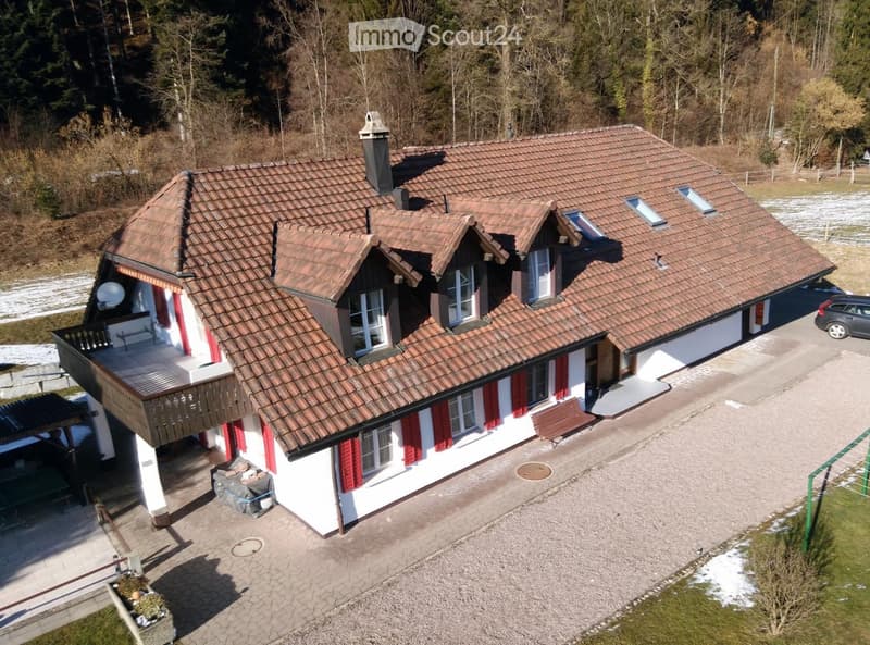 Two-story house with red-tiled roof, white walls, red shutters, balcony, and driveway with parked car.