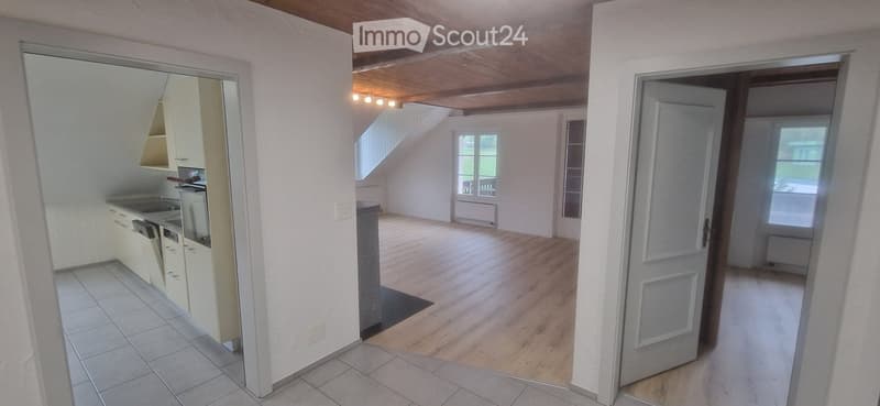 Open-plan living and kitchen area with light wooden flooring, white walls, and modern kitchen units. The kitchen features cabinets, a sink, stove, dishwasher, and refrigerator. A staircase leads to an upper level, and there is a door leading outside.