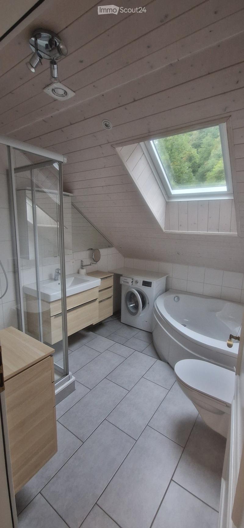 Modern bathroom with a sloped wooden ceiling, skylight window, bathtub, toilet, sink, washing machine, and glass shower enclosure.