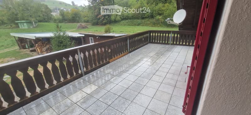 Balcony with tiled floor, wooden railing, and a view of a green landscape with trees and a small structure.