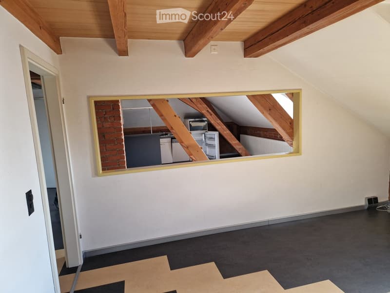 An attic room with white walls, exposed wooden ceiling beams, a window, and a large mirror reflecting the outside area with a refrigerator, microwave, and a brick wall.