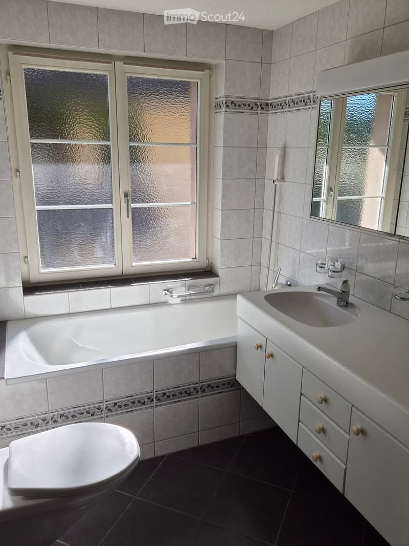 Modern bathroom with a bathtub, toilet, sink, and cabinet. Features grey tiled walls, a dark tiled floor, and a large window with frosted glass.