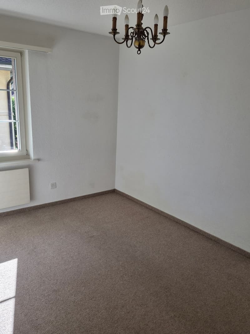 Empty room with beige carpet, white walls, a window with white frame, a radiator, and a chandelier with multiple lights.