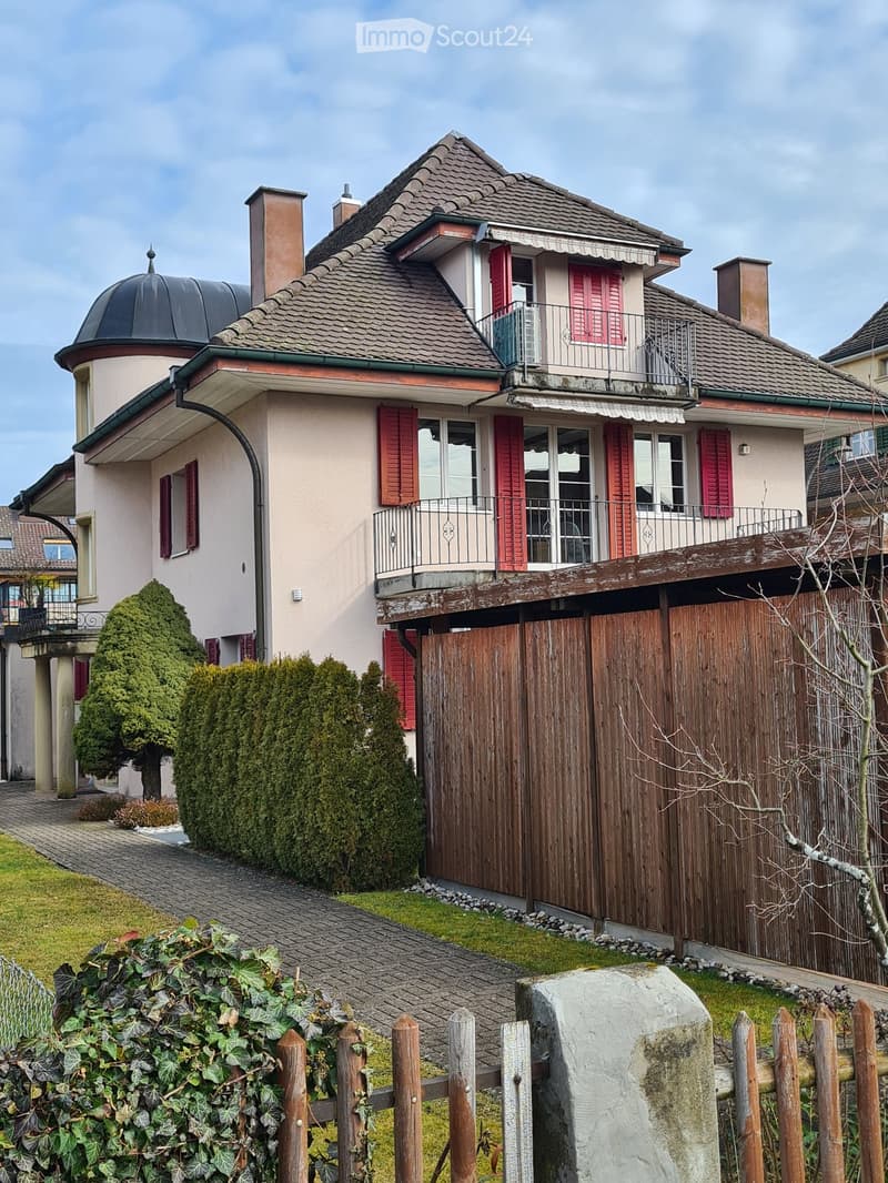 Two-story house with red shutters, a balcony, and a wooden gate. The house features a dome-shaped roof section and is surrounded by greenery and a paved pathway.