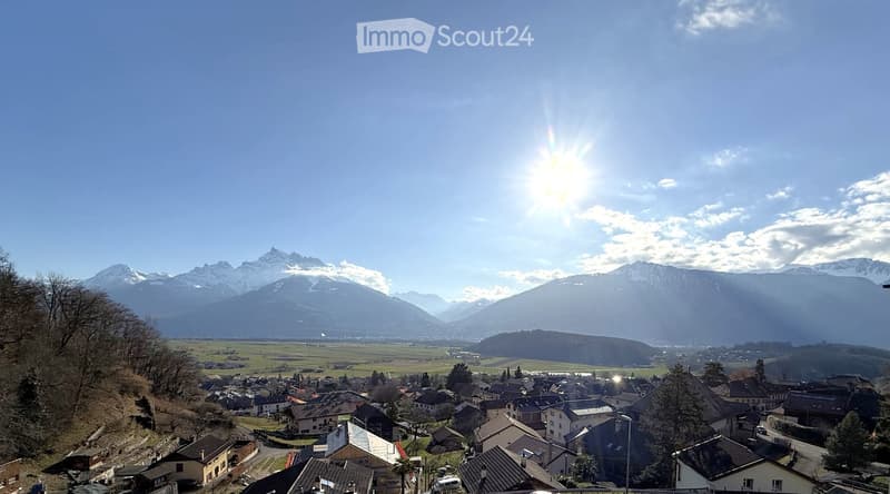 Panoramic view of a village nestled in a valley with snow-capped mountains in the background under a clear blue sky.