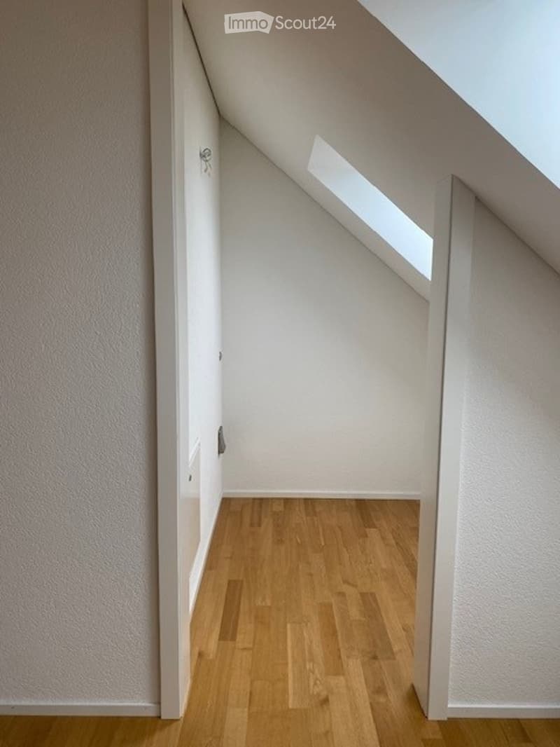 Completely empty attic room with wooden flooring, white walls, and a skylight window.
