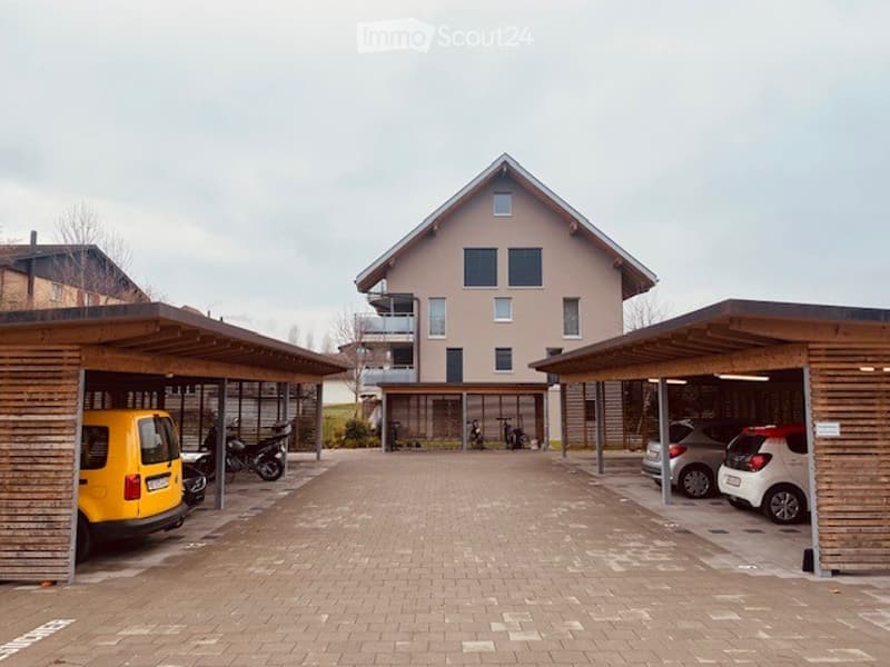 Modern two-story residential building with a covered parking area featuring multiple vehicles, including a yellow van, motorcycles, and several cars.