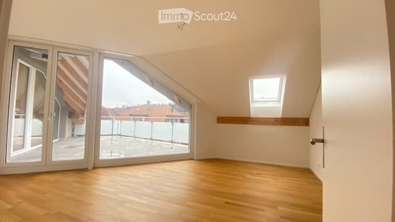 Bright, empty attic room with wooden flooring, large skylight, and large windows opening to a balcony area.