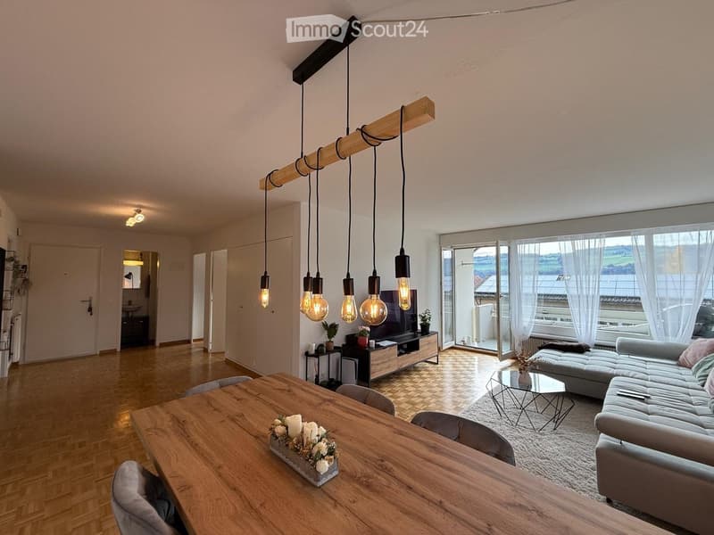 Modern living room with wooden flooring, large windows, a grey sectional sofa, a wooden dining table, pendant lights, and a TV stand.