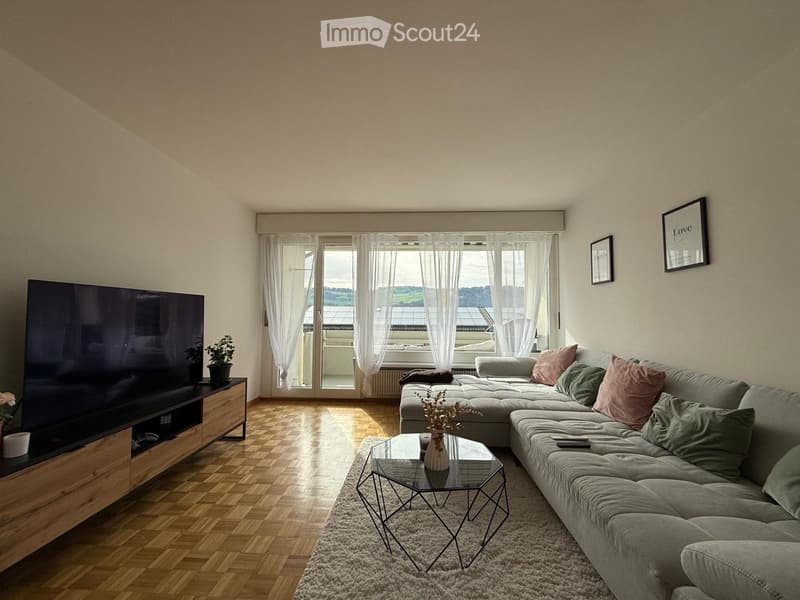 Modern living room with a grey sectional sofa, glass coffee table, flat-screen TV, and large windows with sheer curtains overlooking a view.