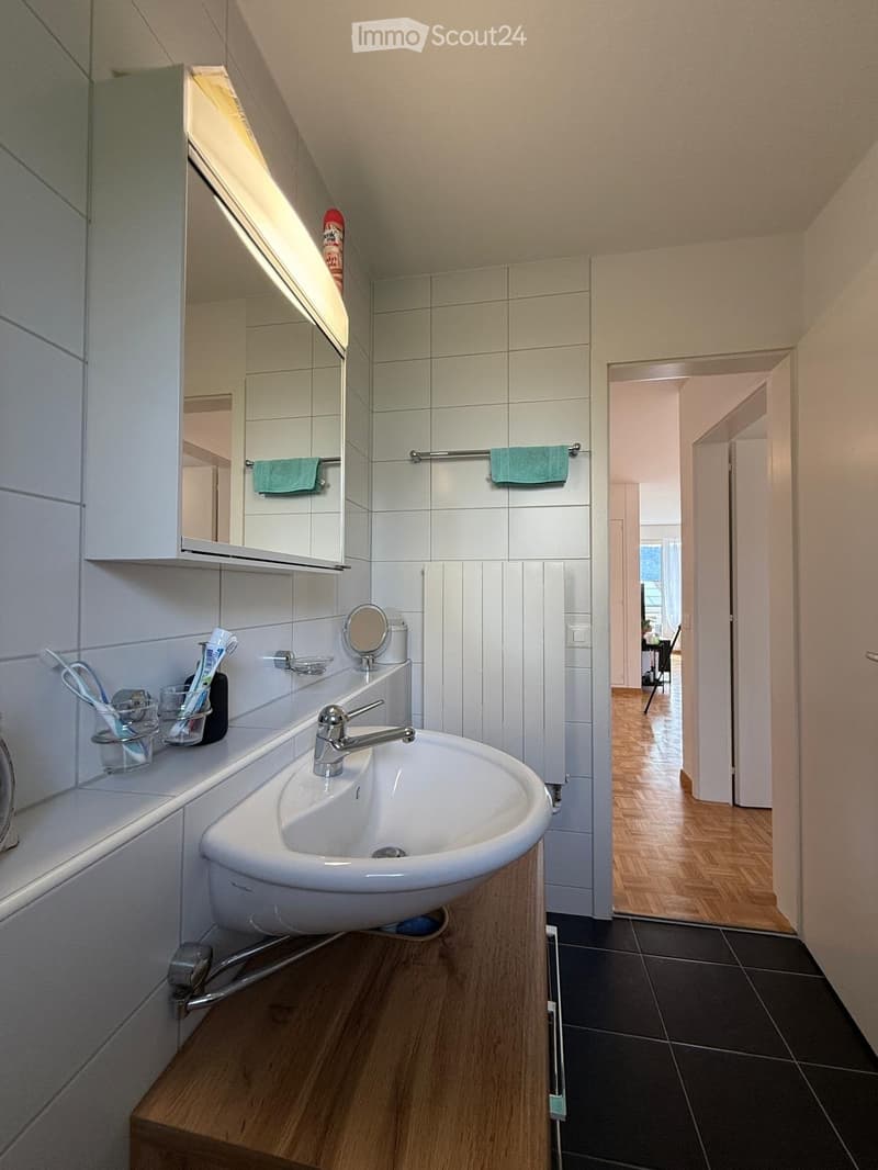 Modern bathroom with a white sink, chrome faucet, mirror, toothbrush holder, and towel rack. The room features white tiles and a wooden floor.