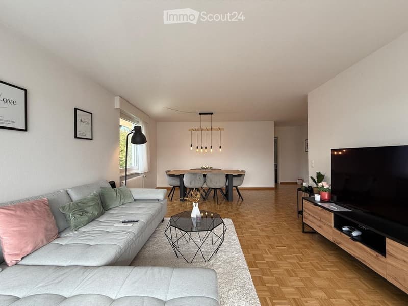Modern living room with a grey sofa, coffee table, dining area, and TV stand. Features wooden flooring, large windows, and decorative elements.