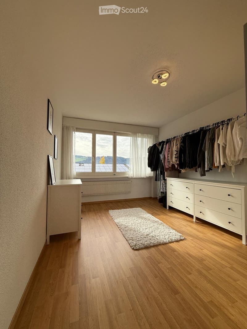 Spacious room with wooden flooring, large windows with white curtains, a white dresser with multiple drawers, a clothing rack with hanging clothes, a small white rug, and picture frames on the wall.