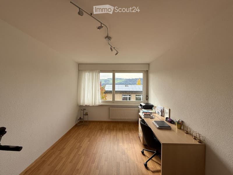 Bright, unfurnished room with wooden flooring, large window with white curtains, desk with chair, laptop, printer, books, picture frames, small table, and stool.
