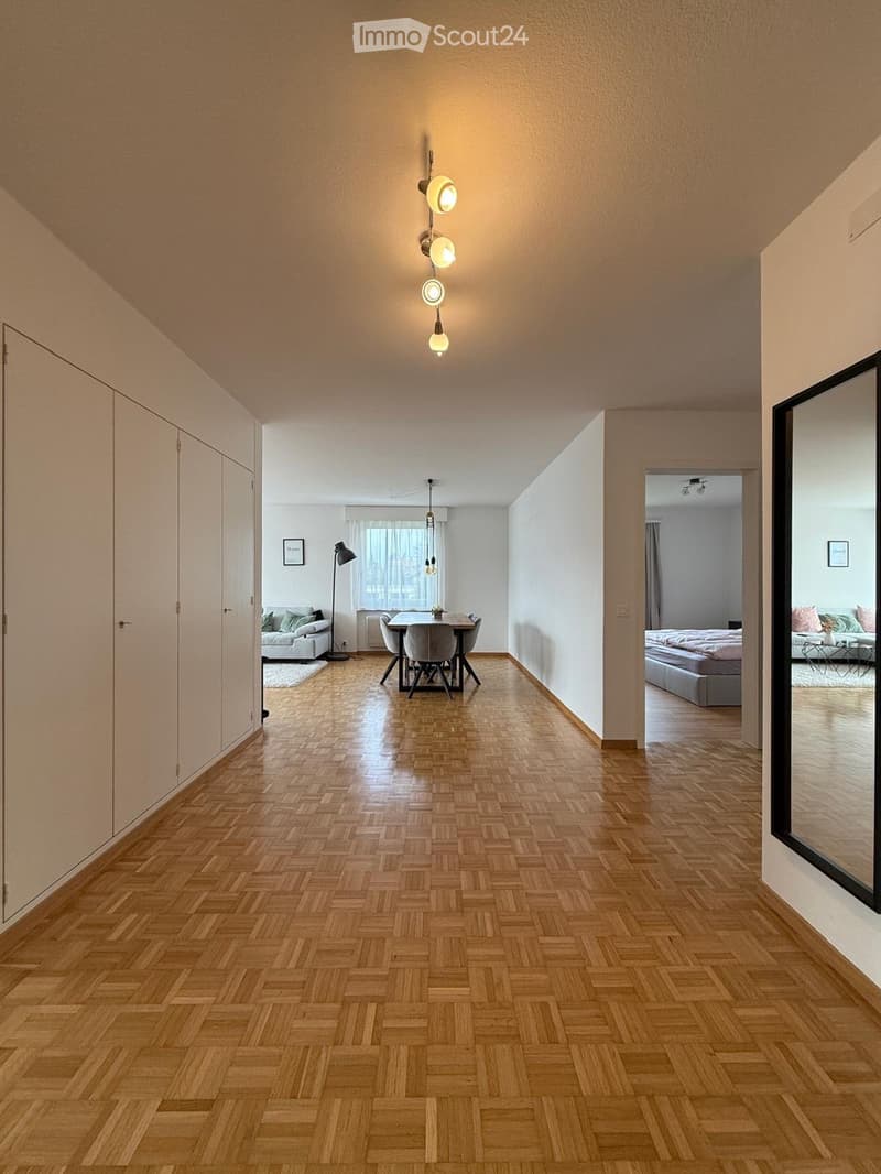 Spacious hallway with herringbone wooden flooring, modern pendant lights, and open access to a living area with a sofa, dining table, and bedroom with a bed.