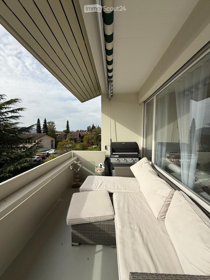 Balcony with a white sofa, two stools, a grill, and a small table, overlooking a residential area with trees and parked cars.