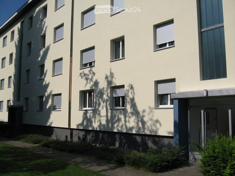Four-story building with light yellow facade, multiple windows with blinds, and a small entrance area with greenery.