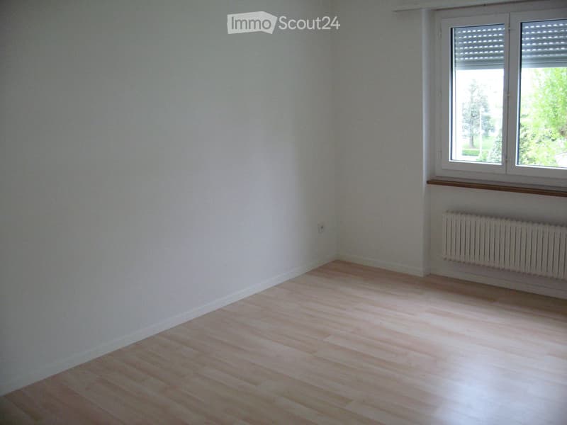 Empty room with light wooden flooring and white walls, featuring a window with blinds and a radiator below it.