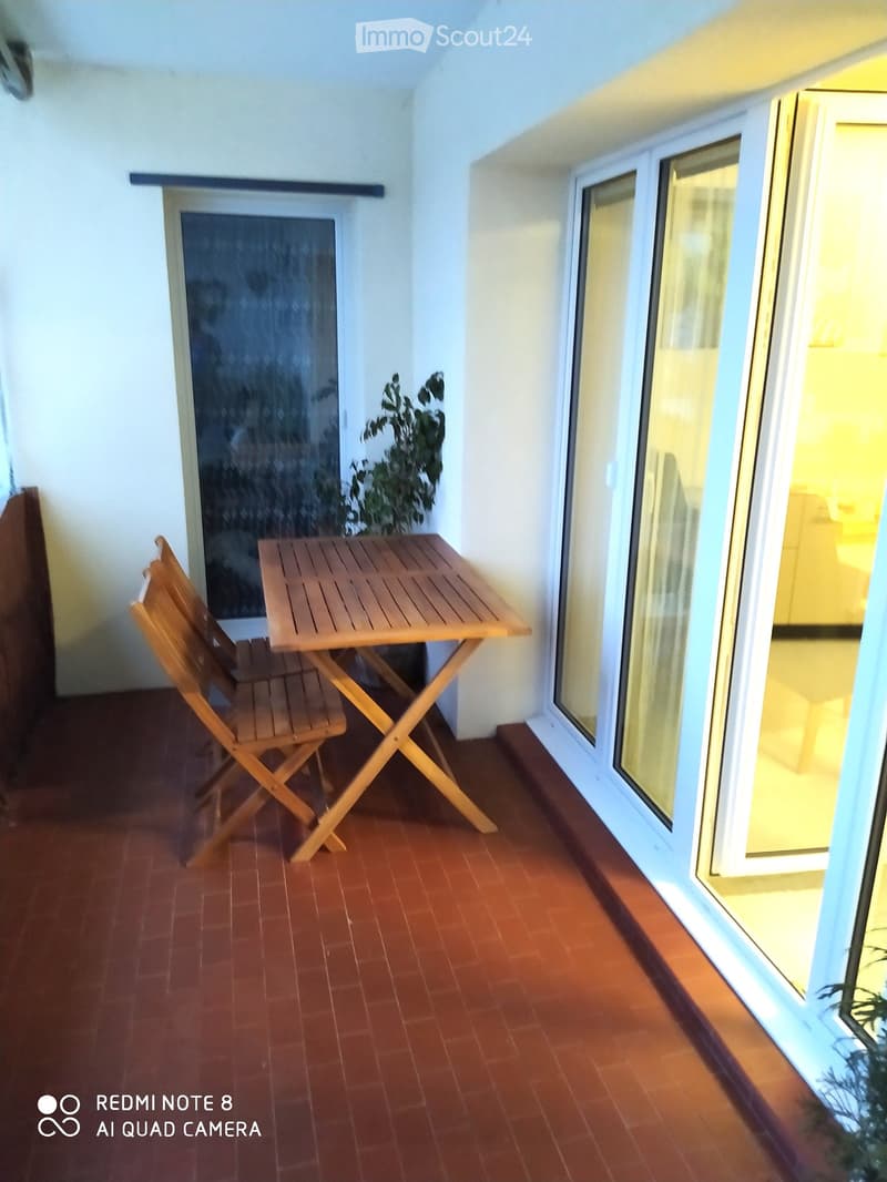 Balcony with wooden table and chairs, terracotta floor, large windows, and indoor view.