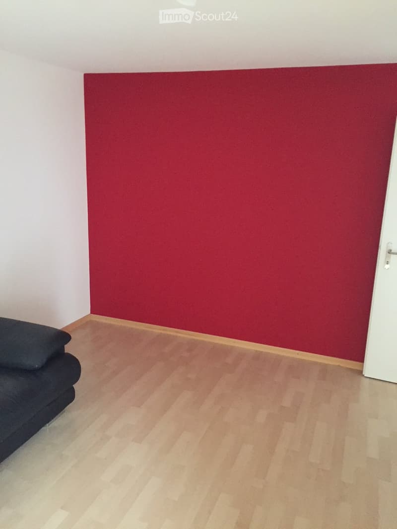 A small, unfurnished room with light wooden flooring and white walls, featuring a dark-colored sofa and a red accent wall.