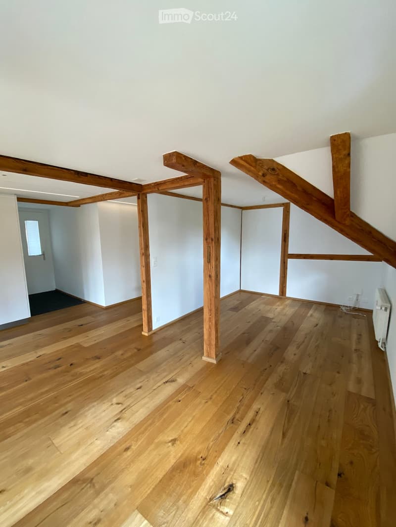 Empty attic space with wooden beams and polished wooden flooring, white walls, and a small radiator visible on the right side.