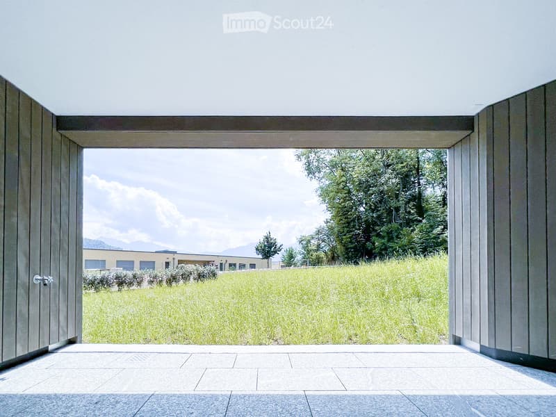 Open corridor with large sliding glass doors leading to a grassy field, distant trees, and mountains under a partly cloudy sky.