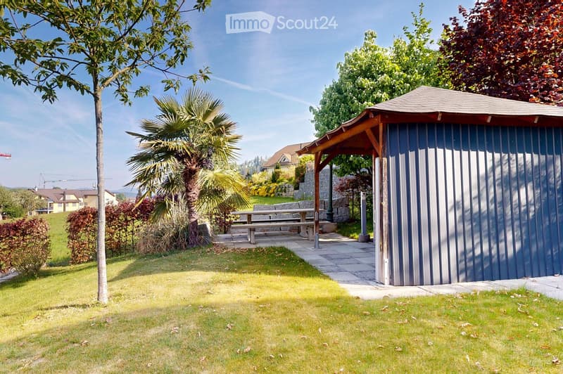 Well-maintained garden with a wooden gazebo, a picnic table, and various trees and shrubs, including a palm tree, under a clear blue sky.