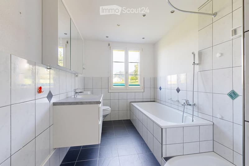 Modern bathroom with white tiles, a bathtub, sink, toilet, and mirrored cabinet. Features a window with a view of greenery outside.