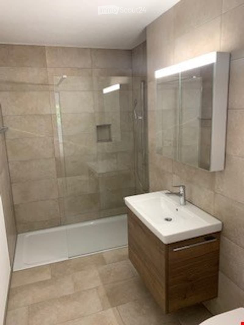 Modern bathroom with a white sink, wooden cabinet, large mirror, and glass-enclosed shower. Beige tiles on walls and floor.