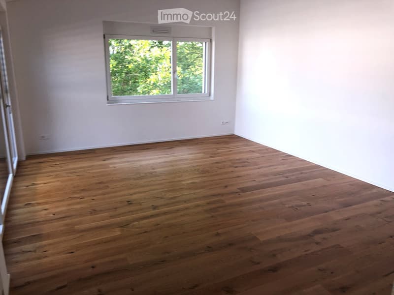 Empty room with wooden flooring and white walls, featuring a large window with a view of greenery.