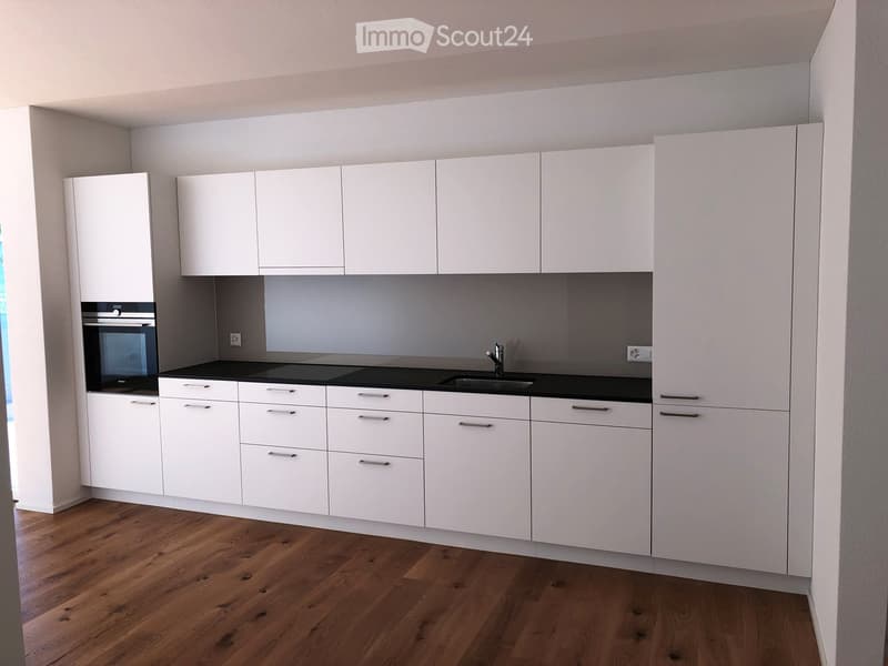 Modern kitchen with white cabinets, black countertops, and a built-in oven. Features a wooden floor and sleek design.