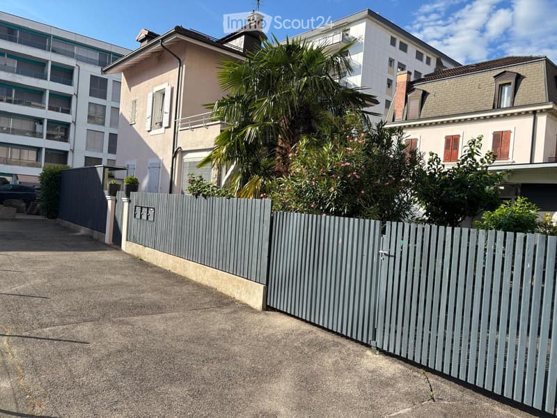A residential area with a multi-story building, a smaller house, and a fenced garden with palm trees and flowering bushes.