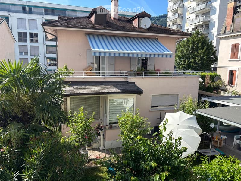 Two-story house with a striped awning, balcony, and garden area with plants and outdoor furniture.