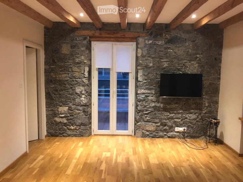Empty room with exposed wooden beams, stone walls, wooden flooring, and a large window with blinds.