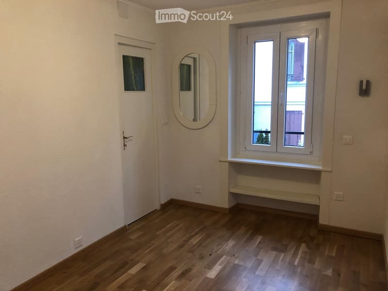 Empty room with wooden flooring, white walls, a door with a small window, a large oval mirror, and a window with a small shelf below it.