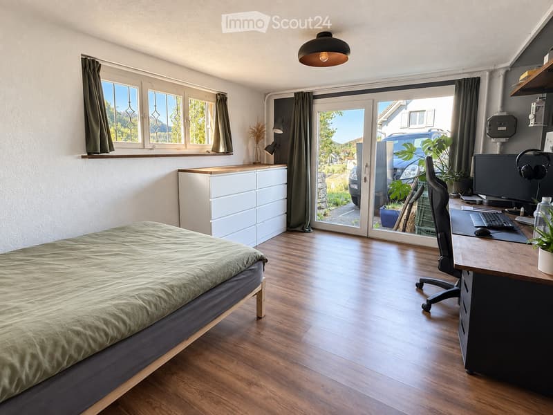 Cozy bedroom with a bed, desk, and large window with a view. Features a wooden floor, modern furniture, and a sliding glass door leading to a patio.