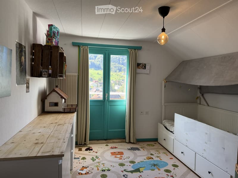 Cozy children's bedroom with a bunk bed, wooden table, shelf, and a view through a large window.