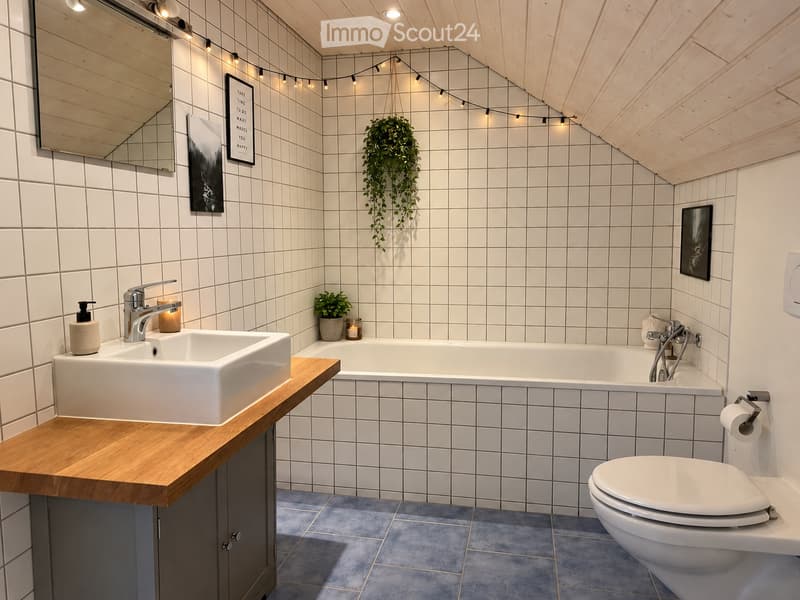 Modern bathroom with white tiles, a wooden vanity, a large sink, a bathtub, and a toilet. Features include a hanging plant, a mirror, and decorative candles.