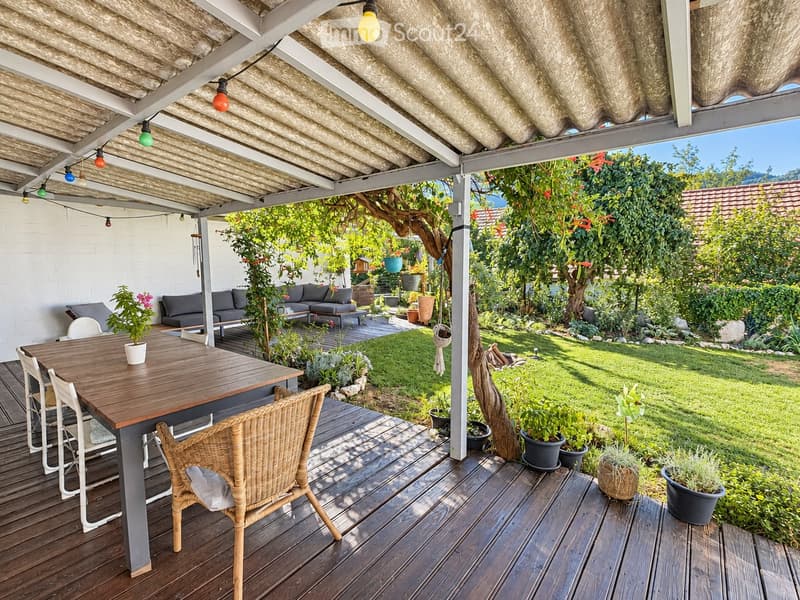 A covered outdoor patio with wooden decking, a dining table with chairs, a sofa, and potted plants, surrounded by a well-maintained garden with grass and trees.