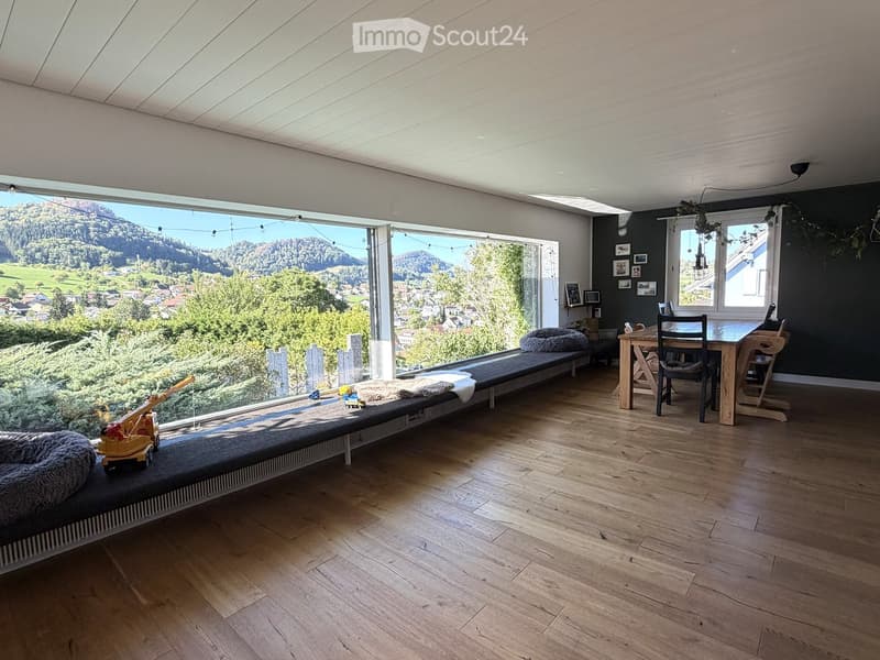 Bright living space with large windows offering a scenic mountain view, featuring a wooden floor, a long sofa, a dining table with chairs, and a toy on the sofa.