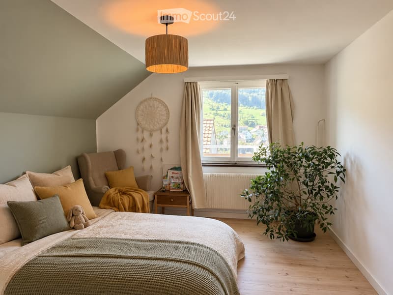 Cozy bedroom with a large window offering a scenic view, featuring a bed with pillows and a blanket, a sofa with cushions, a small table with books, a potted plant, and a dreamcatcher on the wall.