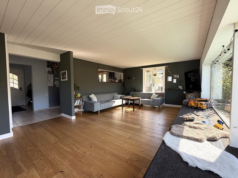 Modern living room with wooden flooring, grey sofa, coffee table, bookshelf, floor lamp, and toys on the floor near the window.