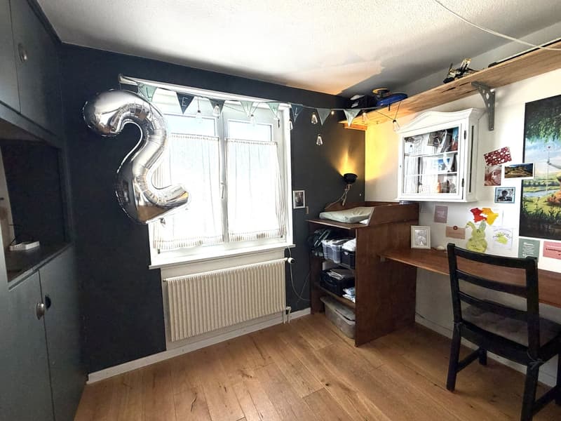 A well-lit room with wooden flooring, a large window with curtains, a desk with a chair, a cabinet, and various decorative items including a balloon and picture frames.