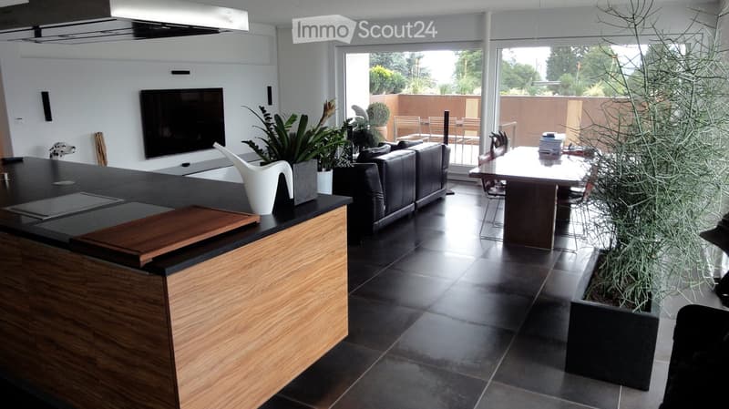 Modern open-plan living and dining area with a dark tiled floor, a black leather sofa, a wooden dining table with chairs, a kitchen island with a wooden cutting board, and a television mounted on the wall. Potted plants and a view of an outdoor patio are
