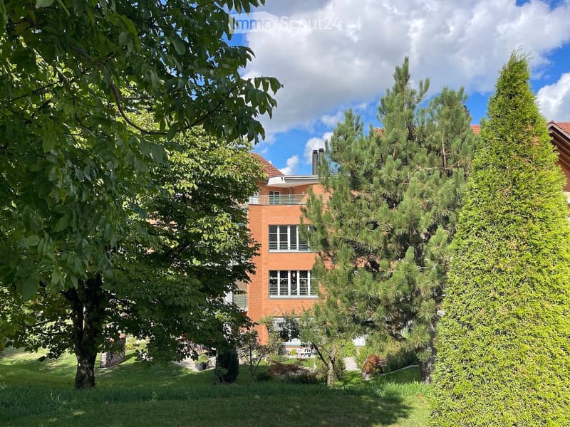 A two-story house with orange brick walls, white-framed windows, and a red-tiled roof, surrounded by lush greenery and trees.