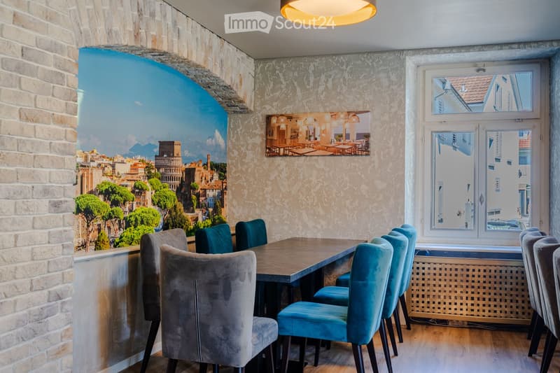A modern dining area with a wooden table and blue upholstered chairs, featuring a large mural of a cityscape and a framed picture of a restaurant on the wall.