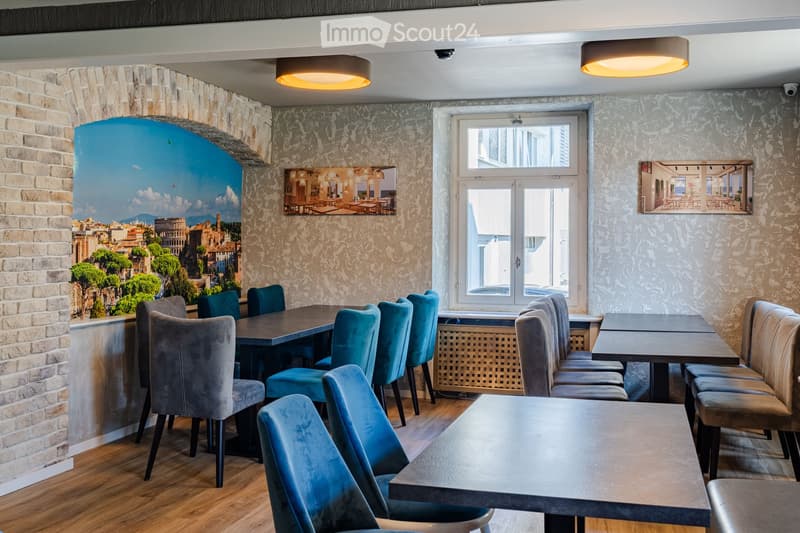 Restaurant interior with wooden tables, blue chairs, brick wall, large window, and artwork.