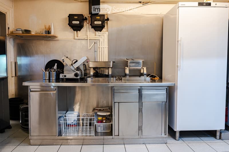 A commercial kitchen with stainless steel appliances, including a refrigerator, sink, cutting machine, and oven. The counter has various kitchen tools and utensils, and there are cabinets and a dish rack below the counter.