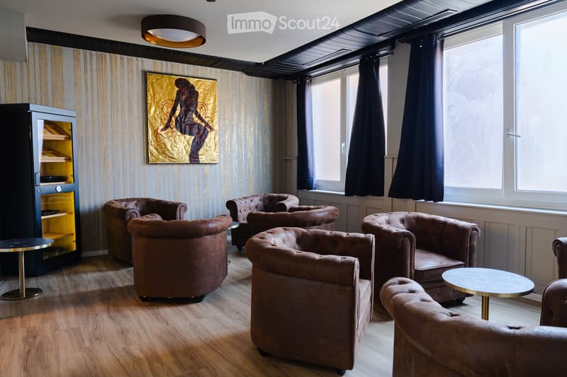 Modern living room with brown leather chairs, small round tables, a cabinet with glass doors, and a large abstract painting on the wall.